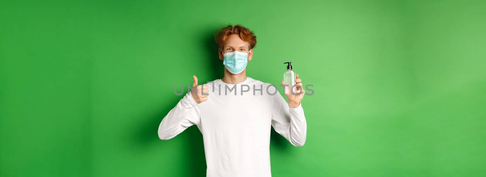 Covid-19, virus and social distancing concept. Smiling young man with red hair, wearing face mask for protection from corona, showing thumb-up and hand sanitizer, green background.
