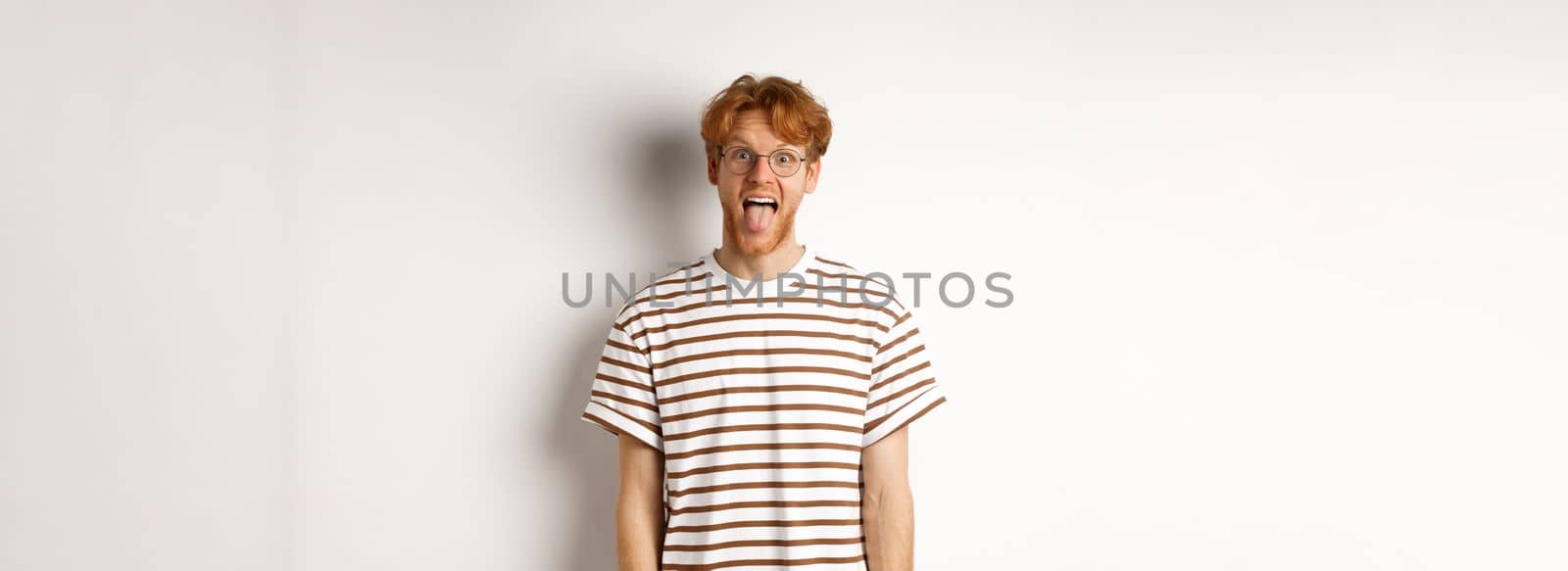 Funny young man with messy red hair and glasses showing tongue, staring at camera, standing over white background.