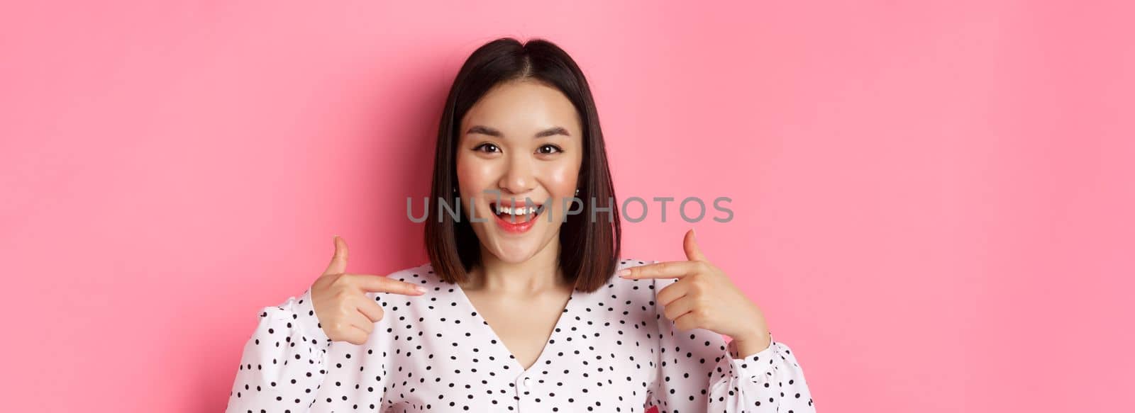 Close-up of happy and confident asian woman pointing at herself, smiling self-assured, standing over pink background.
