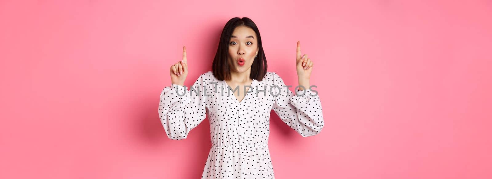 Pretty asian female model in dress pointing fingers up, showing copy space and staring at camera amazed, telling big news, standing over pink background by Benzoix