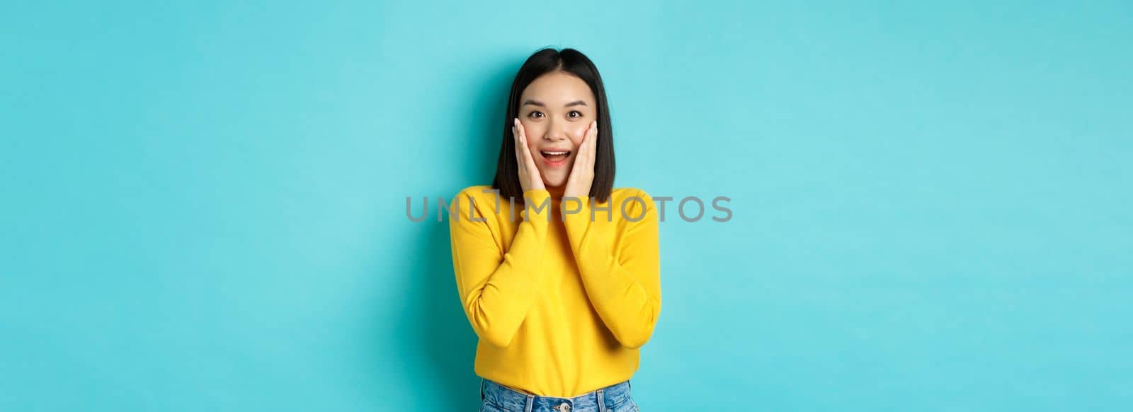 Image of cheerful and surprised asian woman checking out promotion, gasping amazed, standing over blue background.
