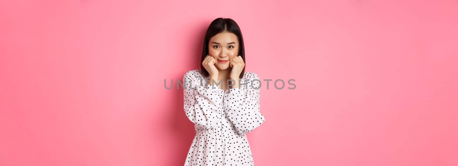 Cute and shy asian girl blushing, touching cheeks and looking at camera silly, standing against pink background by Benzoix