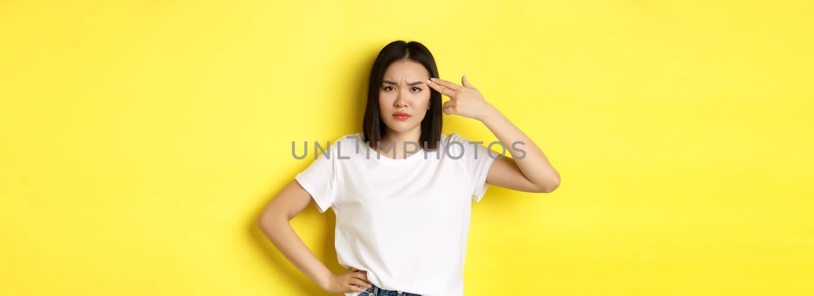 Woman showing kill me please gesture, shooting herself in head with finger gun from boredom, standing upset over yellow background.