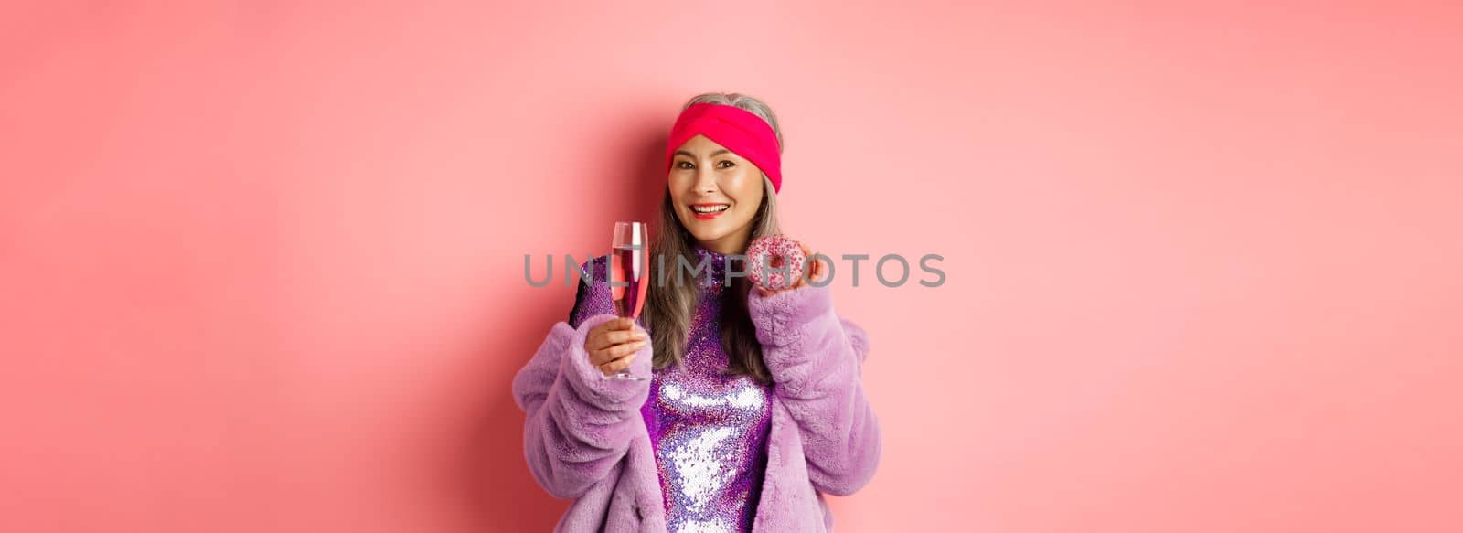 Elegant and stylish asian woman in purple faux fur coat eating donuts and drinking champagne, having fun at party, standing over pink background.