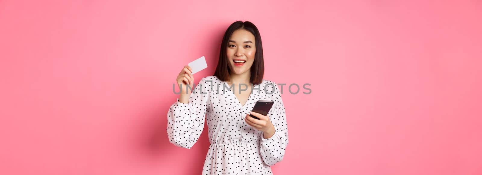 Attractive young asian woman order online, holding credit card and mobile phone, making internet purchase, standing happy over pink background by Benzoix