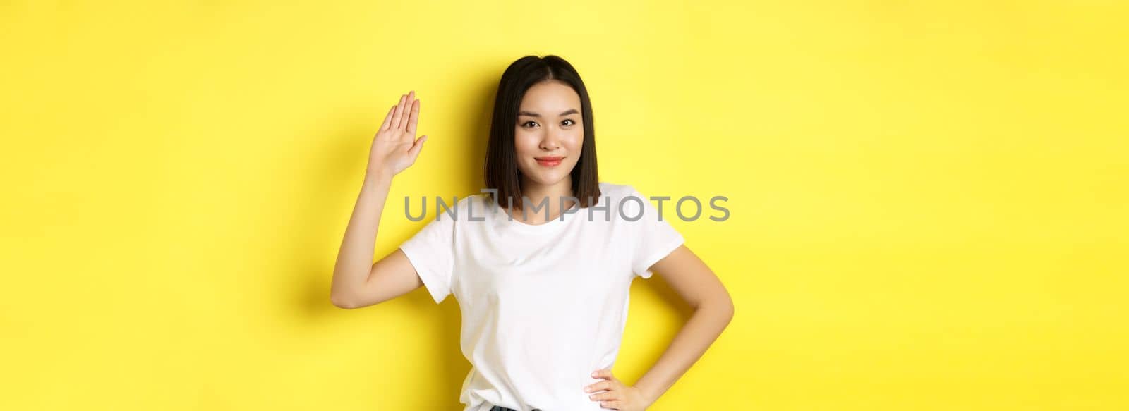 Friendly asian woman in white t-shirt waving hand and saying hello, greeting you, standing over yellow background.