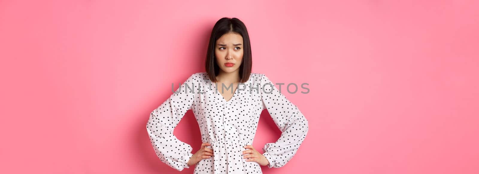 Sad and concerned asian woman having problem, holding hands on waist and looking left with upset face, standing in dress against pink background by Benzoix
