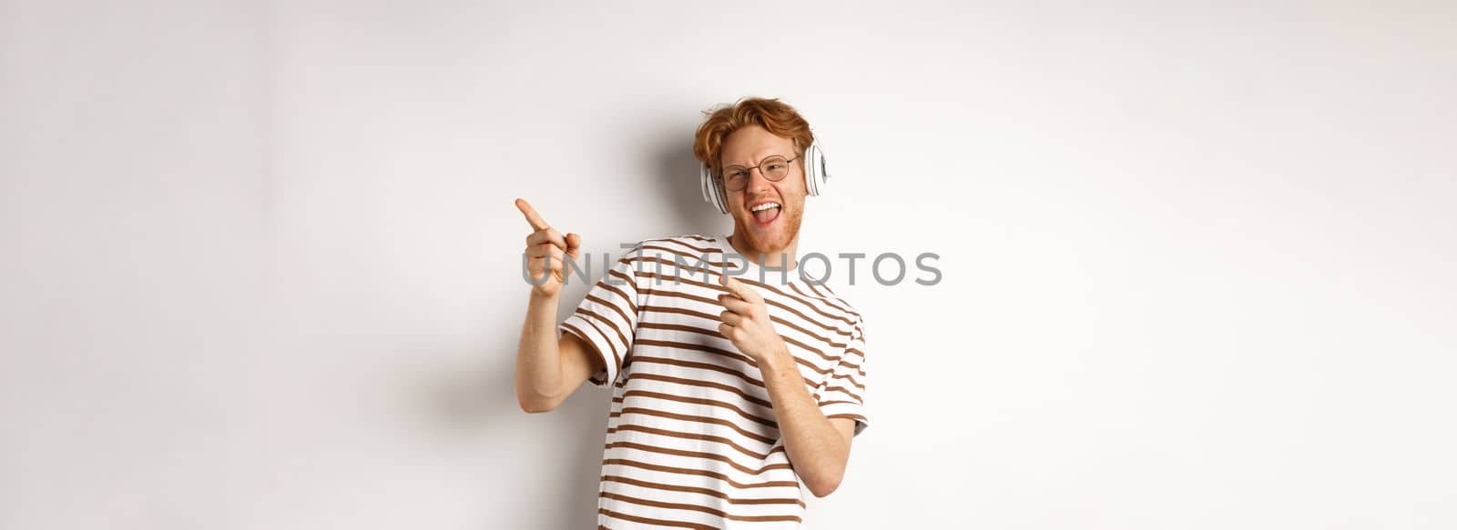 Technology concept. Happy redhead man listening music in headphones and dancing and pointing fingers right, standing over white background.