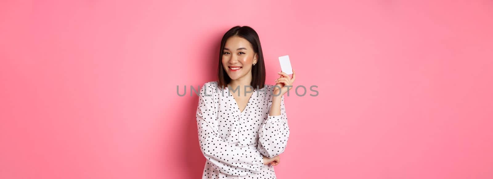 Shopping concept. Confident and happy asian woman holding credit card and smiling satisfied, standing against pink background.