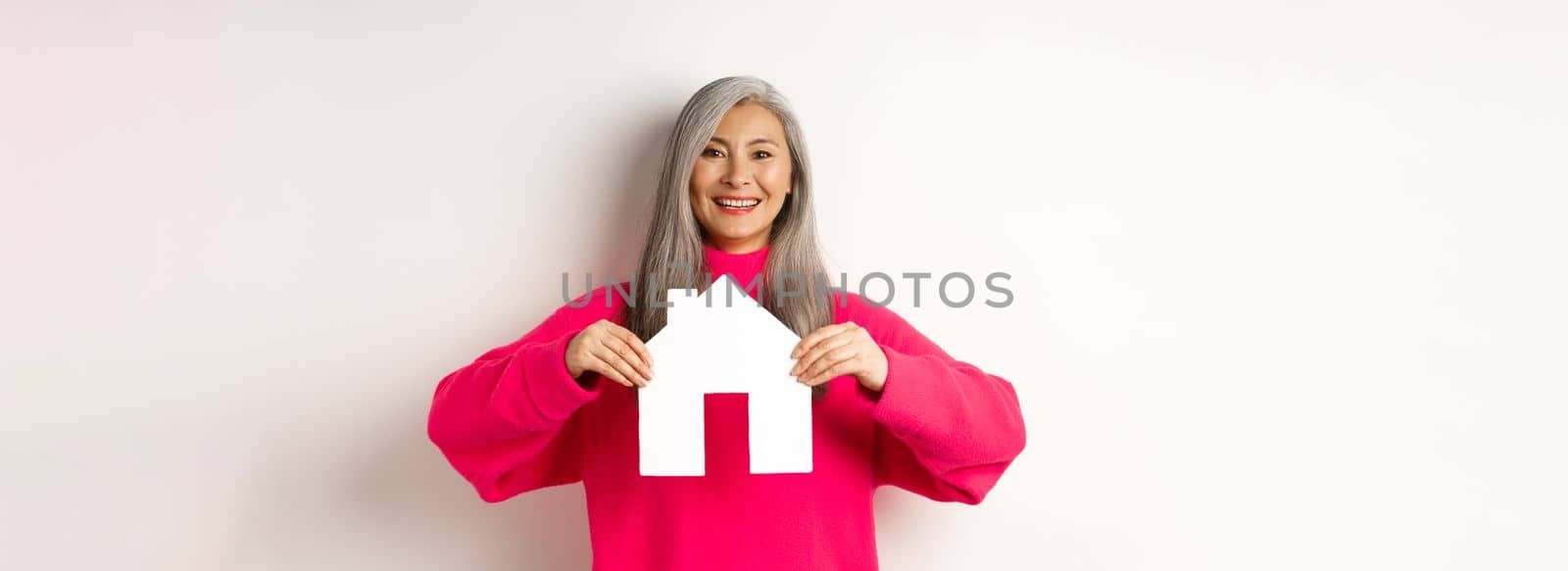 Real estate. Beautiful asian grandmother showing paper house and smiling happy, standing in pink sweater over white background.