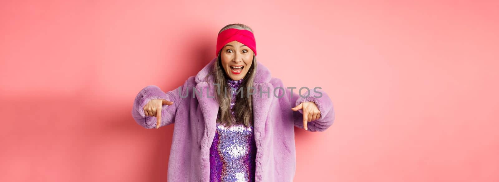 Fashion and shopping concept. Happy korean senior woman pointing fingers down and showing awesome promo deal, wearing trendy purple fur coat and headband, pink background.