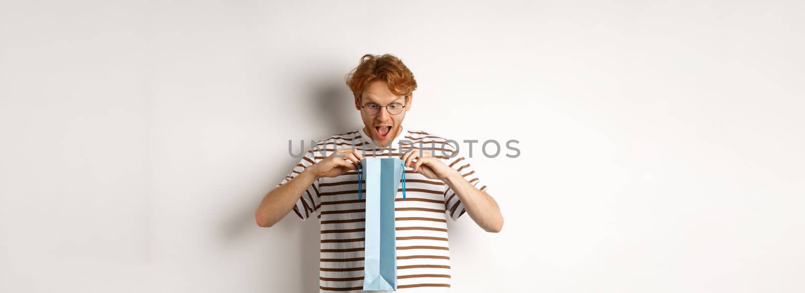 Surprised boyfriend open shopping bag with valentines day gift, staring inside with amazed face, standing over white background.