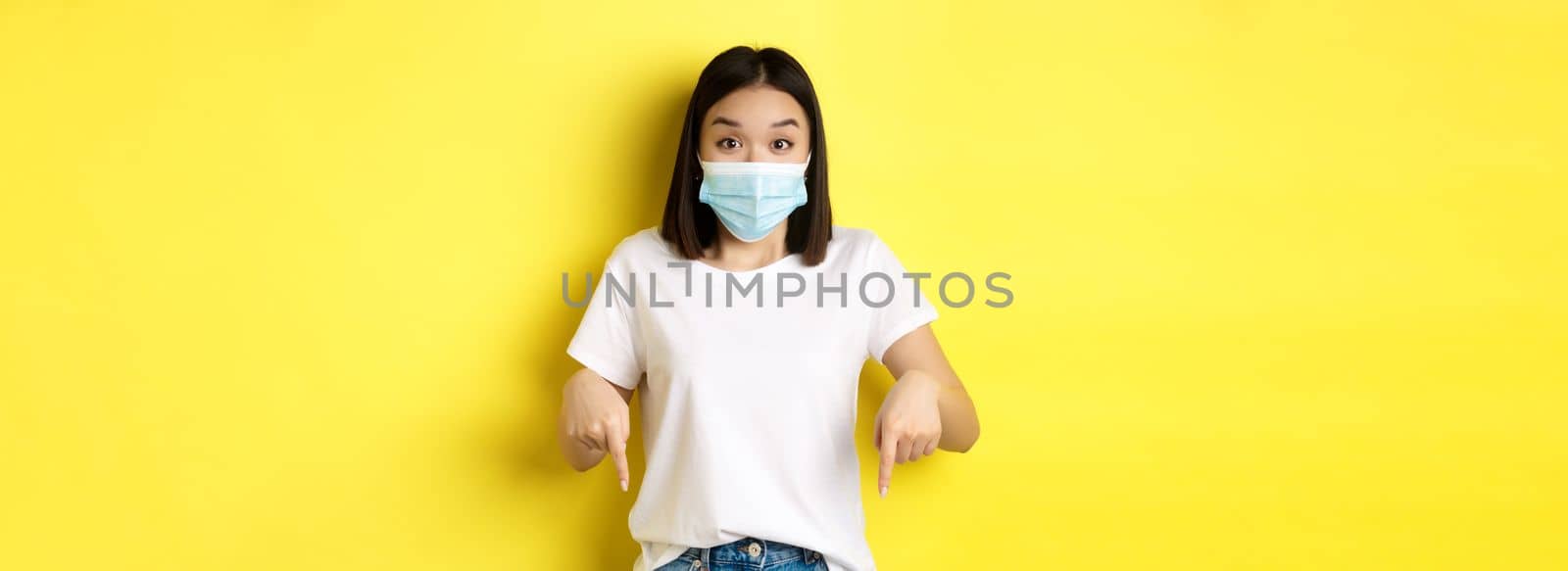 Covid-19, pandemic and social distancing concept. Amazed asian woman in medical mask, showing advertisement, pointing right and smiling, yellow background.