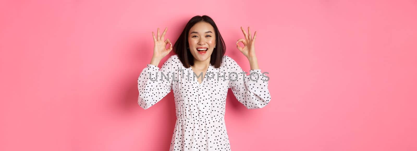 Pretty young asian woman in dress showing okay sign, praising and showing approval, looking satisfied, standing against pink background by Benzoix