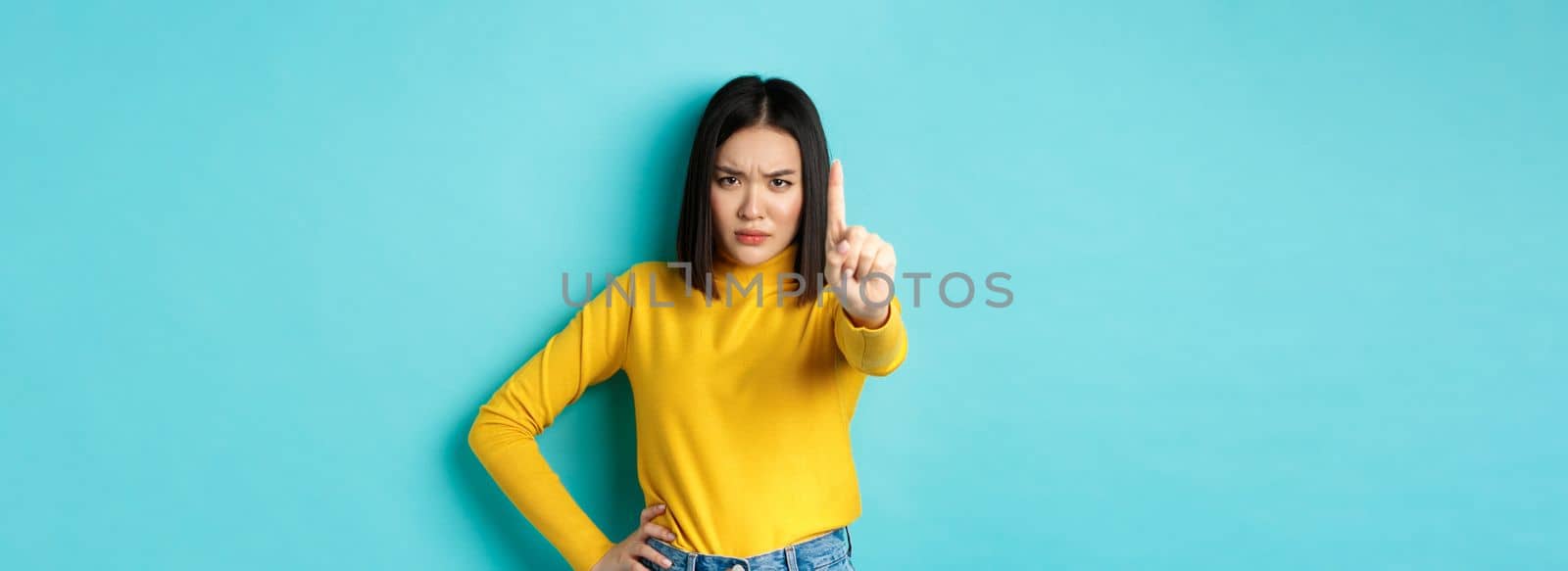 Confident and serious woman tell no, showing extended finger to stop and prohibit something bad, frowning and looking at camera self-assured, standing over blue background by Benzoix
