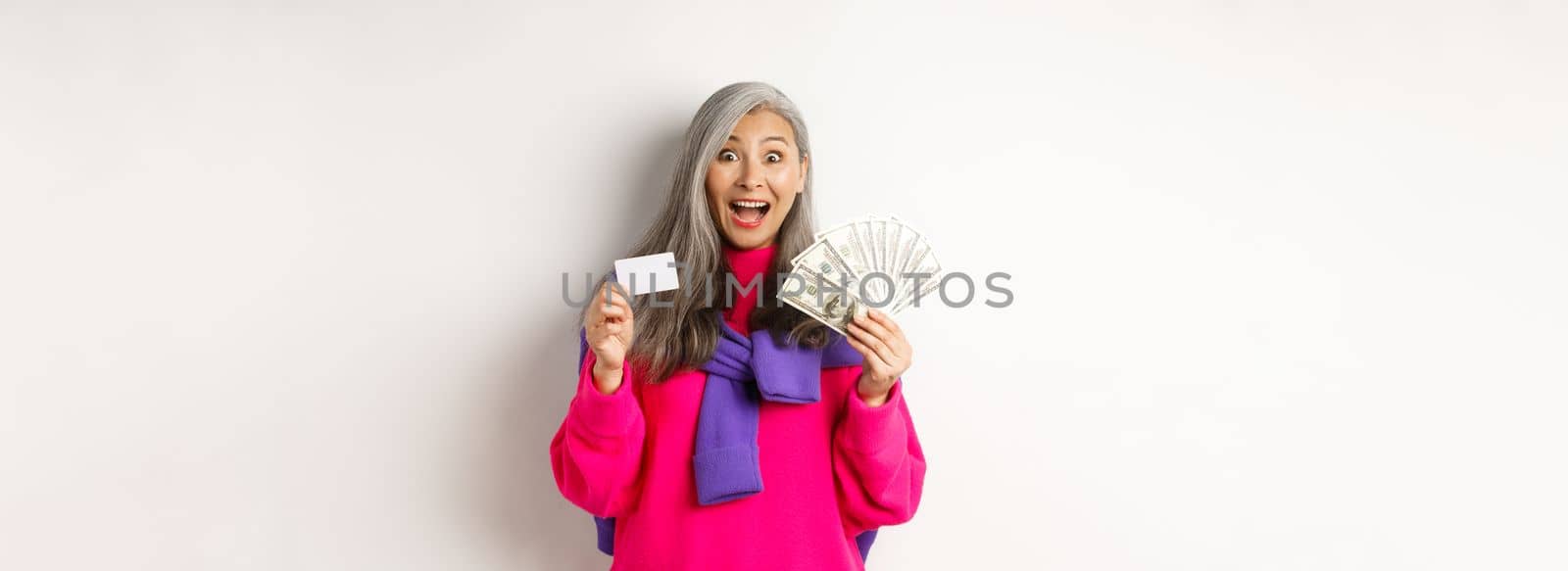 Shopping concept. Amazed asian granny showing plastic credit card and money dollars, want to buy something, standing over white background.