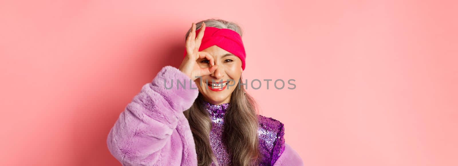 Fashion. Close-up of fashionable asian senior woman smiling, showing OK sign over eye and looking happy at camera, standing over pink background.