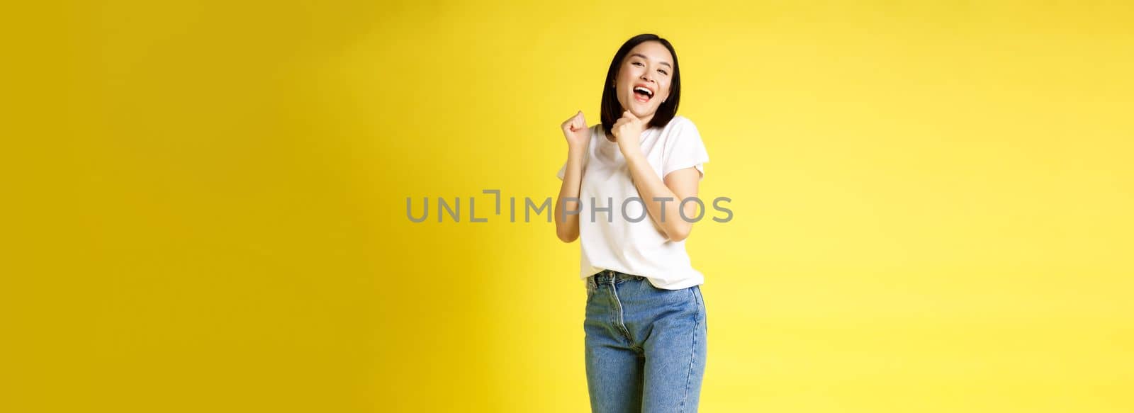 Full size shot of happy asian woman dancing and jumping from happiness, winning and celebrating victory, posing over yellow background.