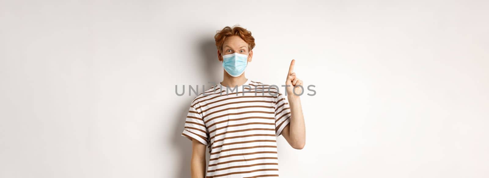 Covid, virus and social distancing concept. Surprised redhead man in face mask showing advertisement on top, pointing finger up and look amazed, white background.
