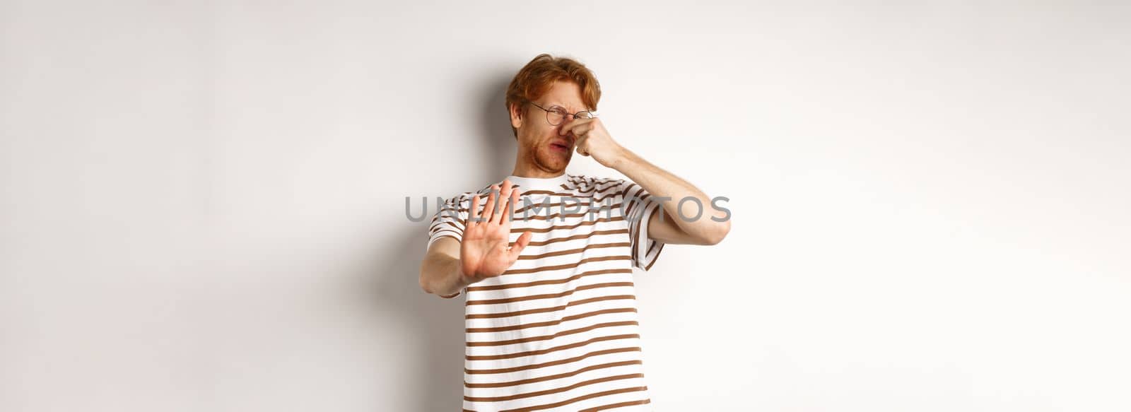 Young man with red hair staring at camera and shut nose from bad smell, looking disgusted and refusing, white background by Benzoix