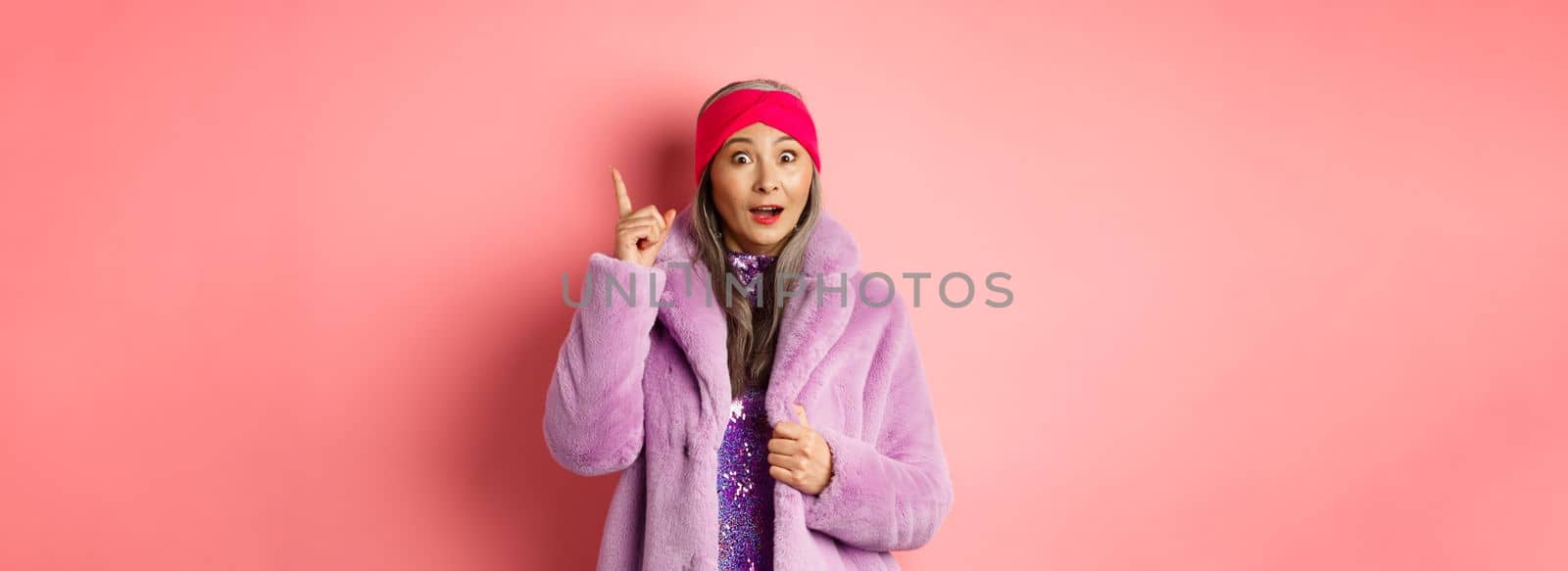 Fashion and shopping concept. Smiling elderly woman having an idea. Stylish old lady in purple fake fur coat raising index finger, suggesting plan, standing excited on pink background.