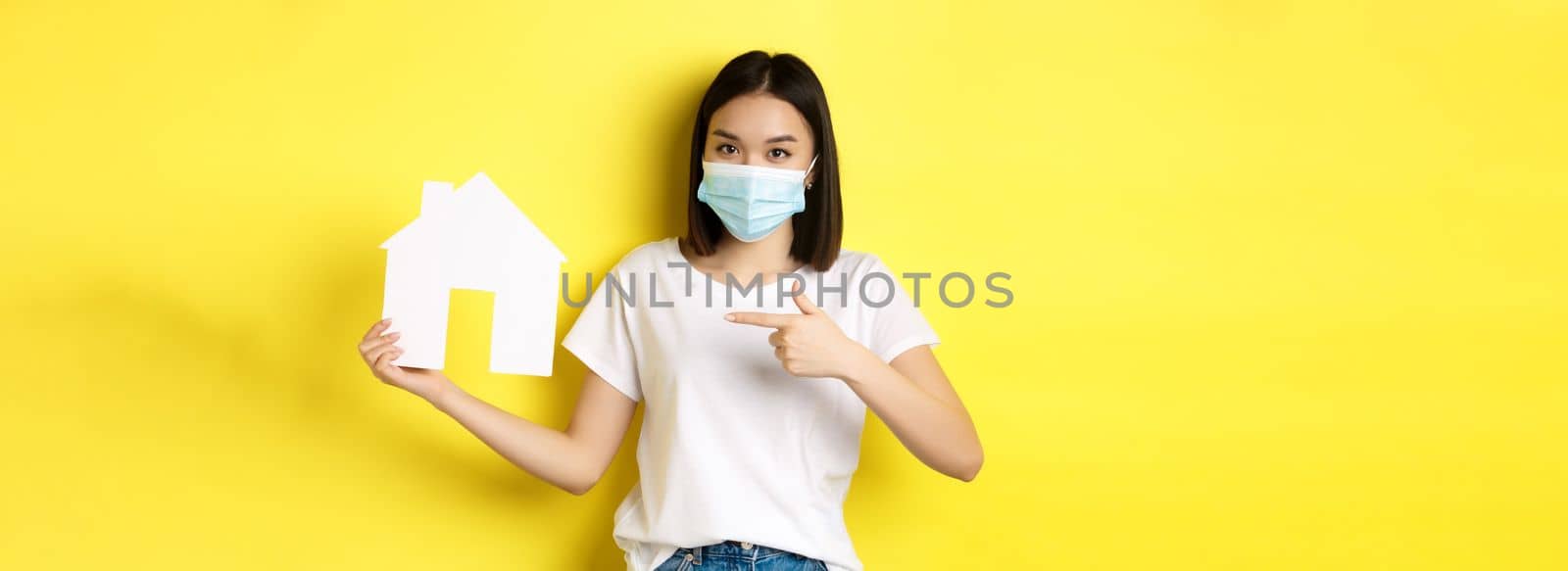 Covid-19, pandemic and real estate concept. Cheerful asian woman smiling in medical mask, showing paper house cutout, recommend agency for buying property by Benzoix