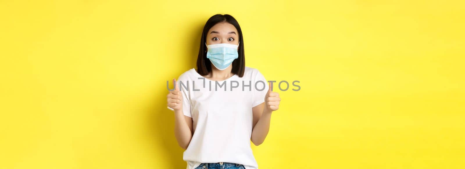 Covid-19, quarantine and social distancing concept. Cheerful asian woman in medical mask and white t-shirt showing thumbs up in approval, praise good deal, yellow background.