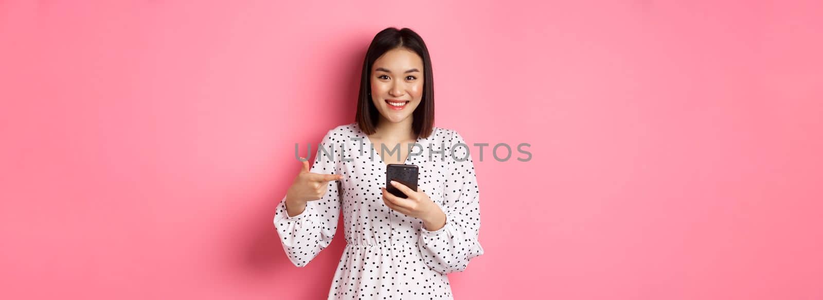 Beautiful young asian woman in romantic dress using smartphone, smiling and pointing at mobile phone, standing over pink background.