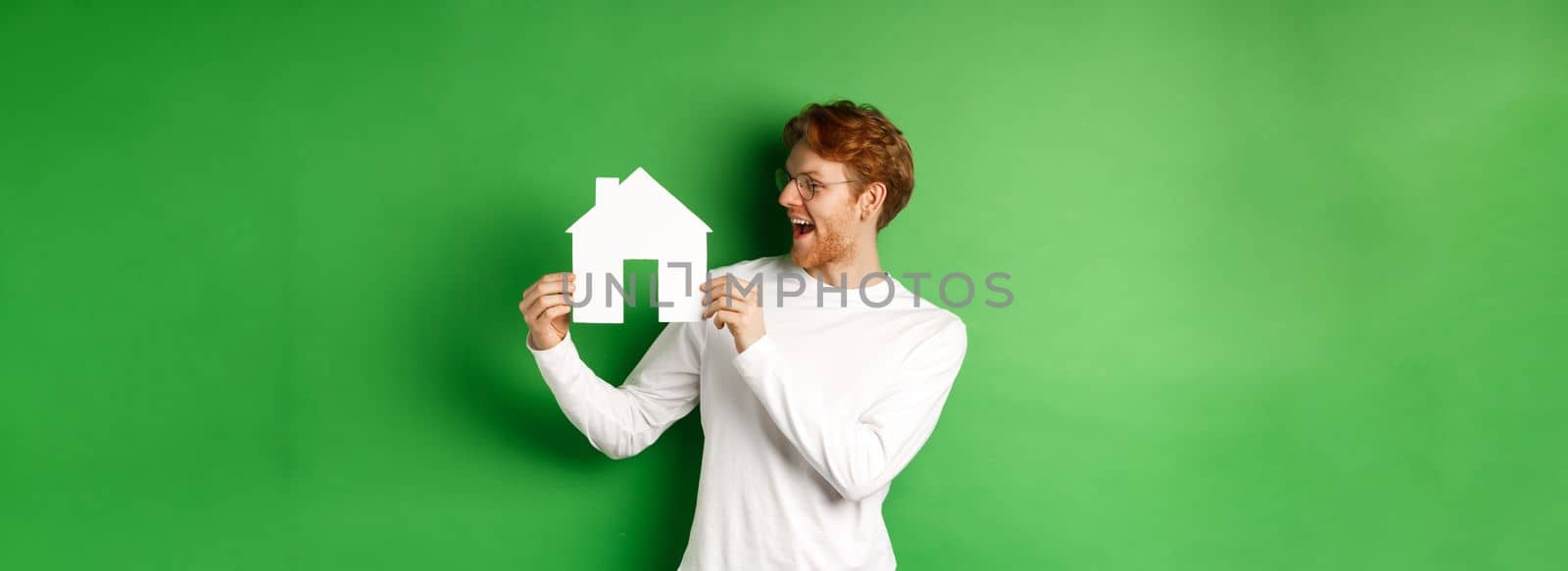 Real estate. Cheerful caucasian man with red hair, looking at paper house cutout and smiling amazed, buying property, standing over green background.