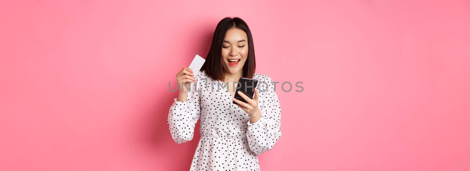 Attractive young asian woman order online, holding credit card and mobile phone, making internet purchase, standing happy over pink background by Benzoix