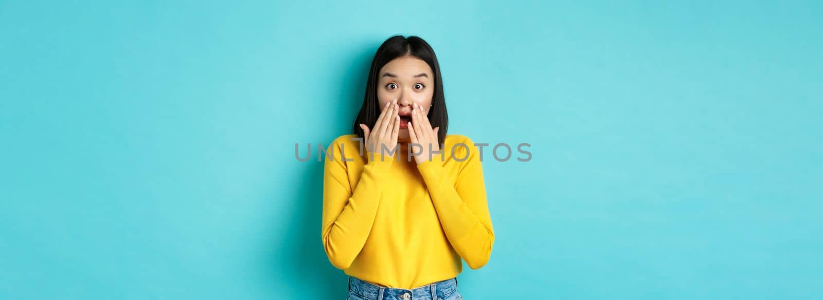 Portrait of impressed korean girl saying wow, gasping and staring amazed at camera, standing in yellow pullover over blue background.