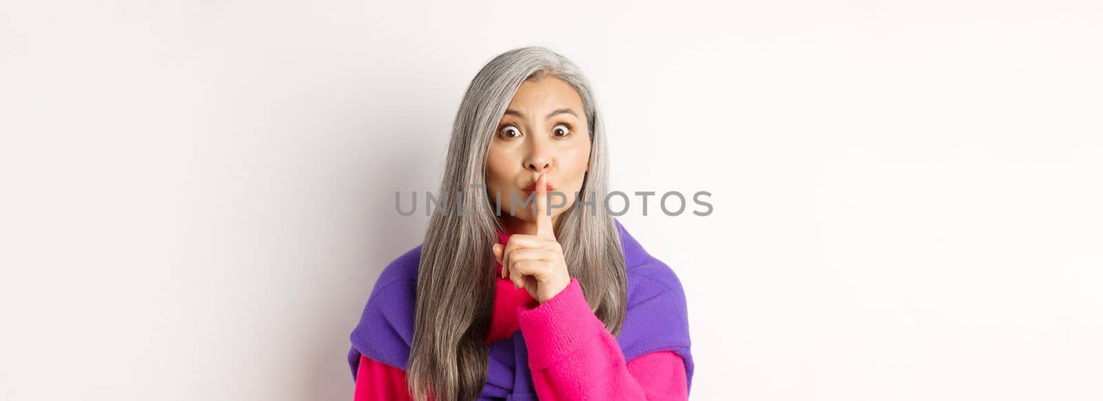 Close-up of stylish senior asian woman in hipster outfit telling hush, shushing at looking at camera, show taboo gesture, standing over white background.