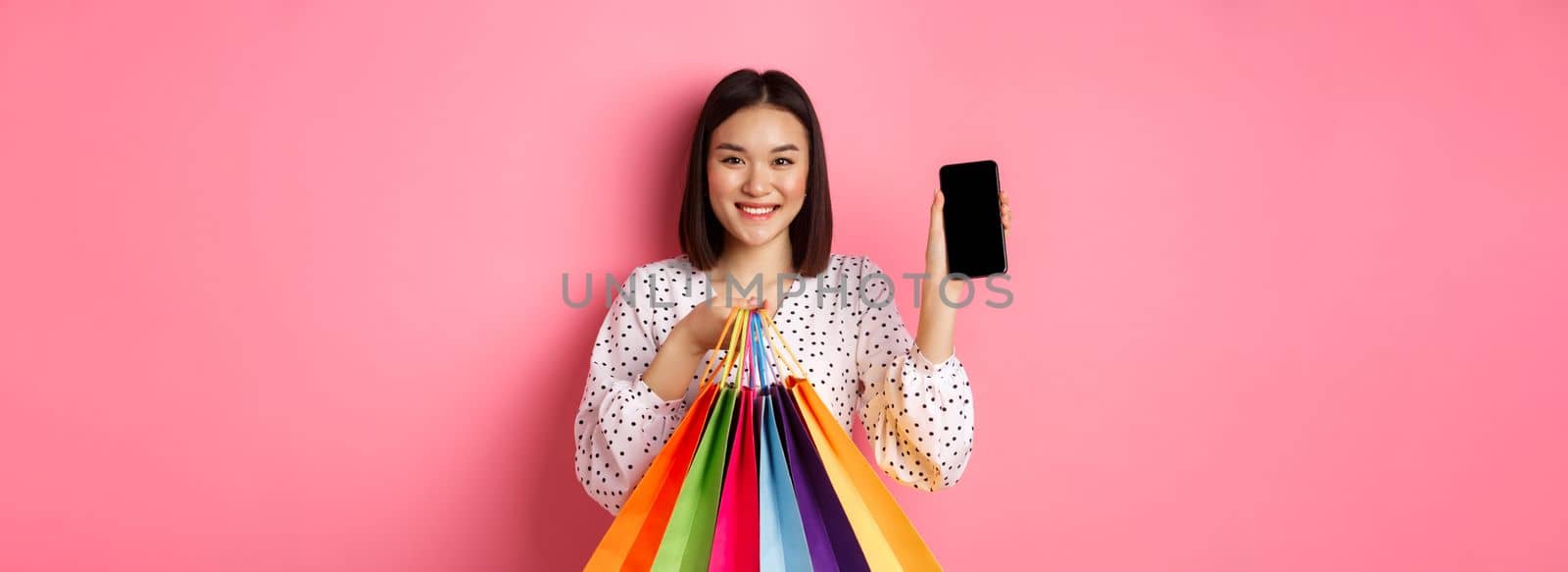 Attractive asian woman showing smartphone app and shopping bags, buying online via application, standing over pink background.