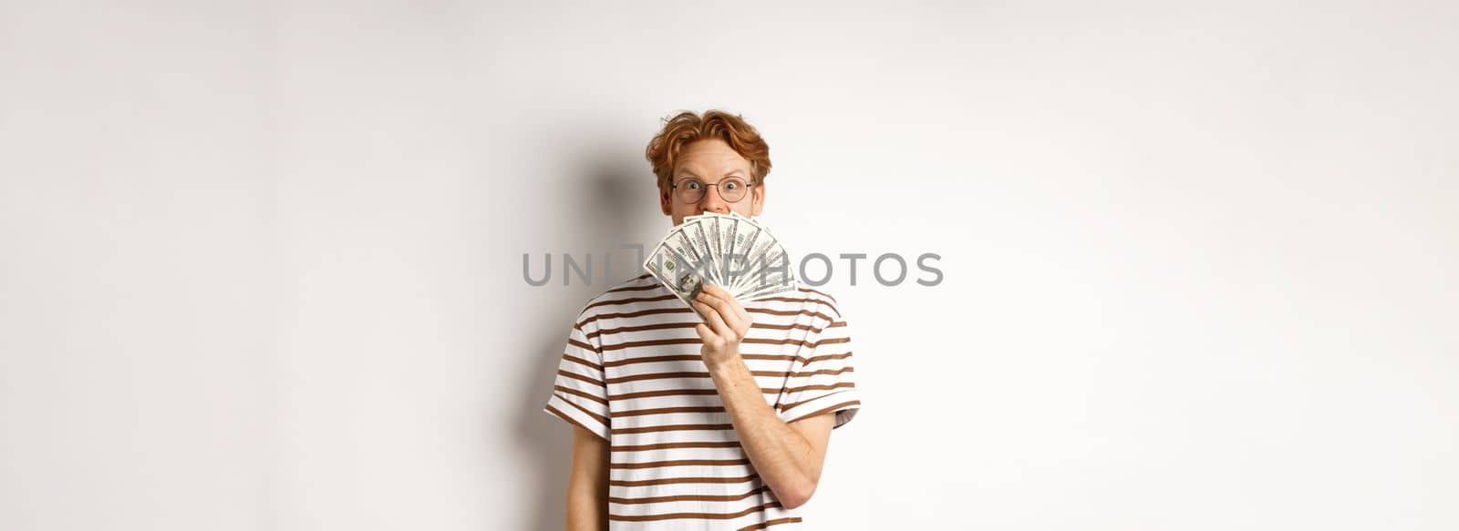 Shopping and finance concept. Lucky redhead guy winning, showing prize money and smiling happy at camera, standing in glasses and t-shirt over white background.