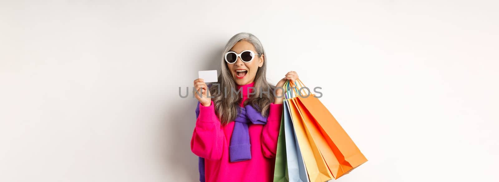 Stylish asian grandmother in sunglasses going shopping on holiday sale, holding paper bags and plastic credit card, standing over white background.