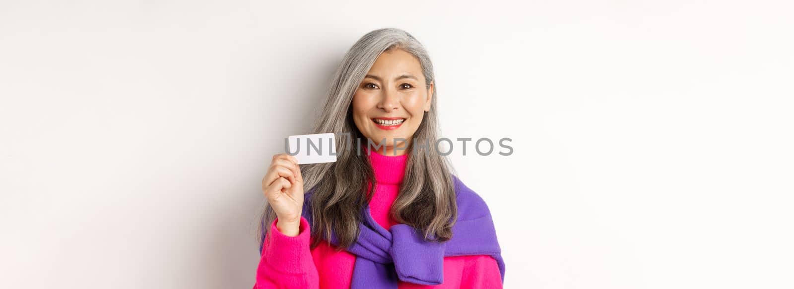 Close-up of beautiful asian senior woman in stylish clothes, smiling and showing plastic credit card of bank, standing over white background.