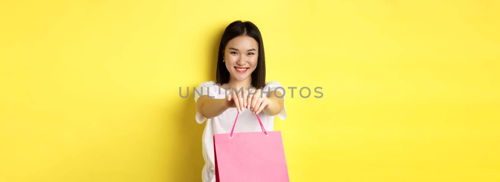 Cute asian girl giving you gift, stretch out hand with pink shopping bag and smiling, congratulating with holiday, standing over yellow background.