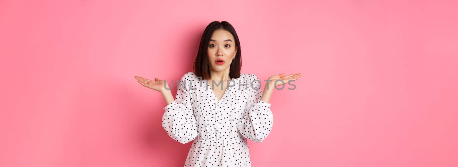 Confused and clueless asian girl shrugging, staring at camera puzzled, dont know what to do, spread hands sideways, standing over pink background.