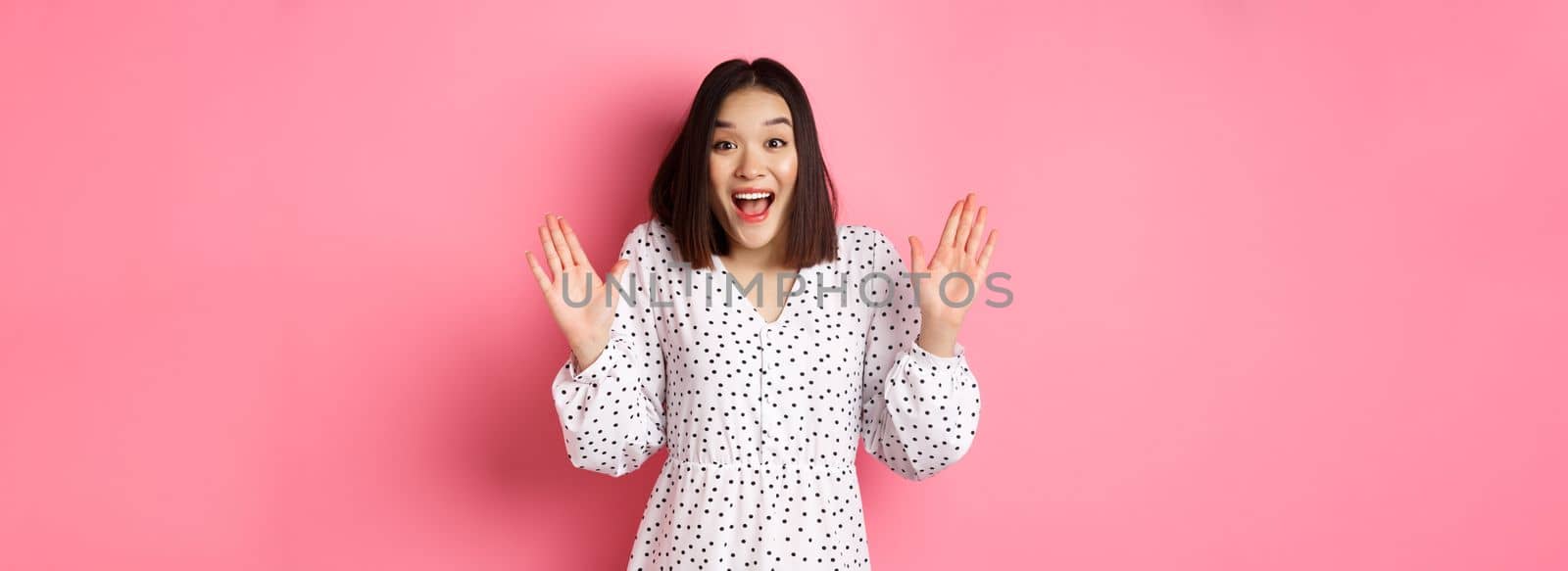 Happy and surprised asian girl receive surprise, looking glad hearing good news, standing amazed in dress against pink background by Benzoix