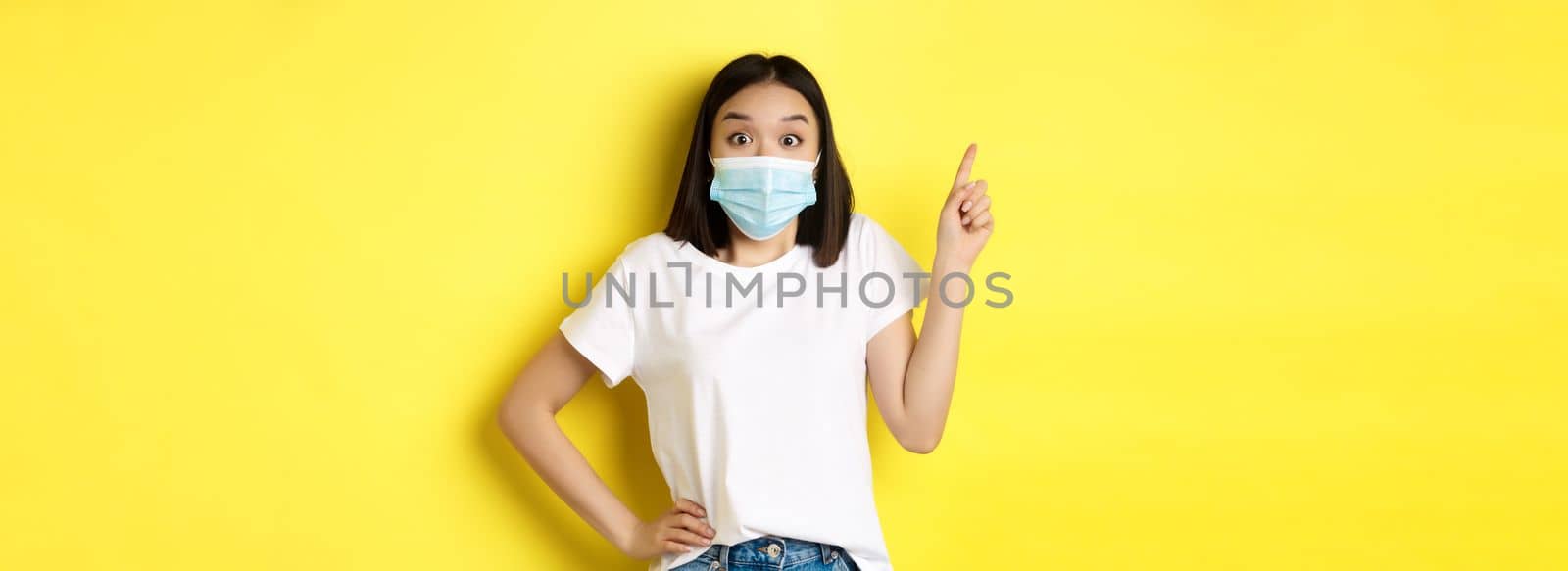 Covid, health care and pandemic concept. Asian female model in medical mask and white t-shirt pointing finger at upper left corner logo, showing promotion, yellow background.