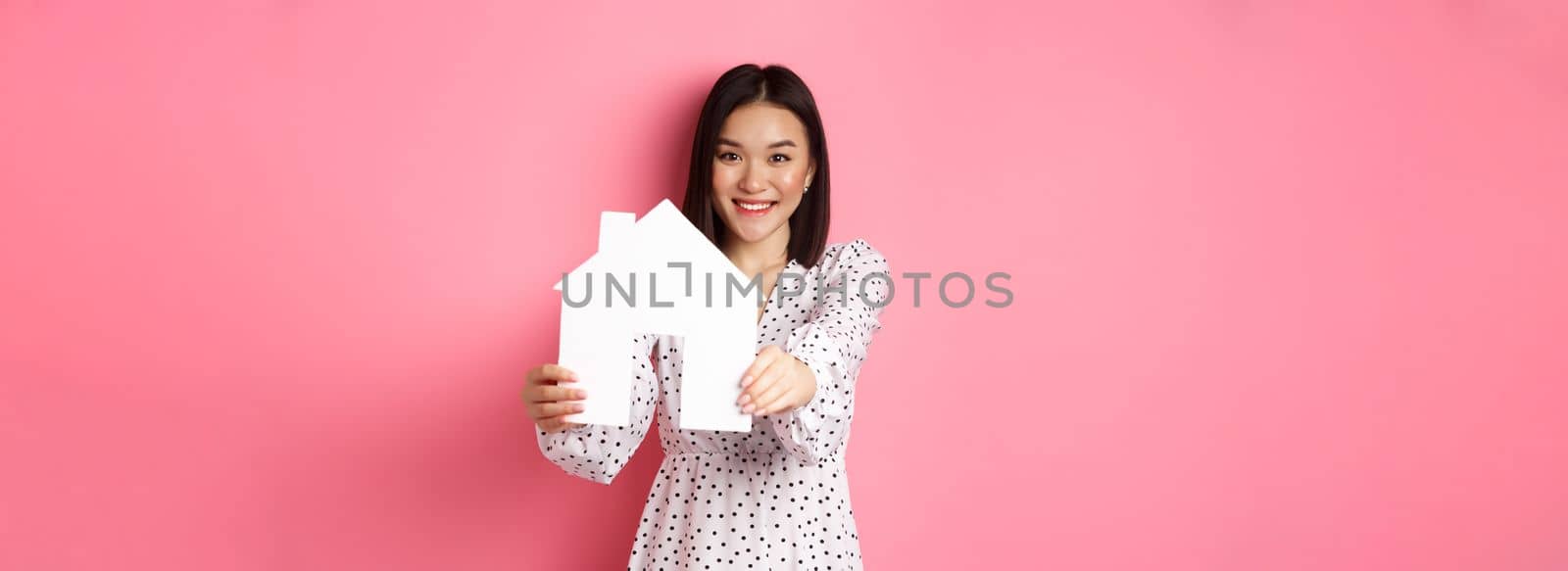 Real estate. Adult asian woman searching for home, holding house model and smiling, promo of broker company, standing over pink background by Benzoix