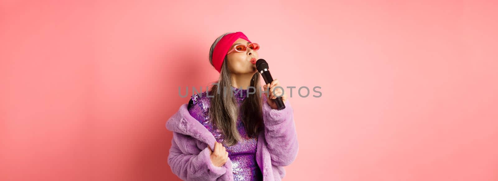 Stylish asian senior woman singing song, performing karaoke with microphone, standing in party outfit and faux fur coat against pink background.
