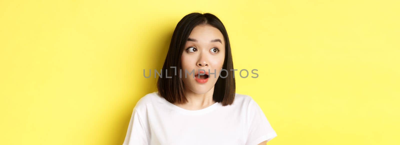 Close up of surprised asian girl drop jaw, gasping and looking left at logo, standing over yellow background.