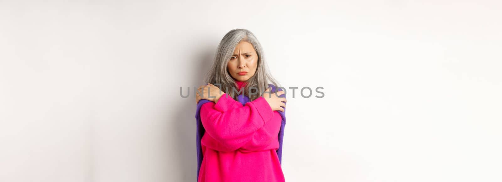Defensive and offended asian middle-aged woman hugging body, comforting herself and looking angry at camera, frowning insulted, standing over white background.