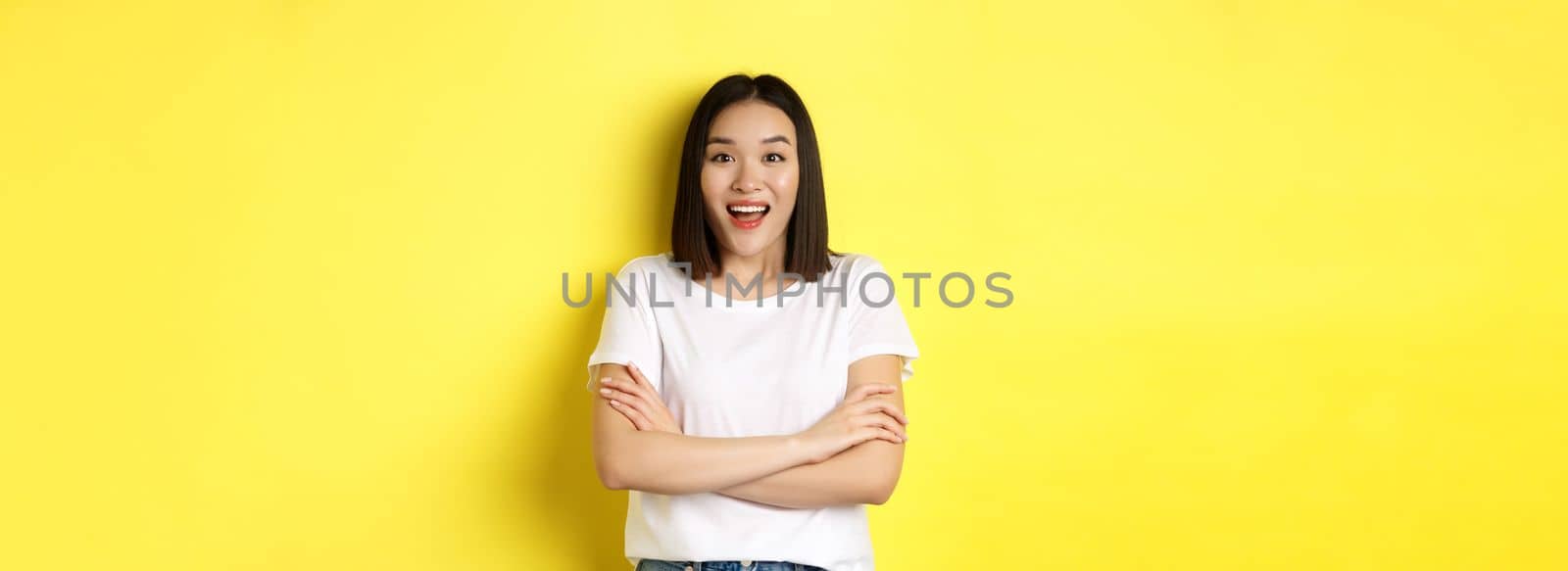 Beauty and fashion concept. Happy asian woman in white t-shirt, cross arms on chest, looking amazed at camera, standing over yellow background.