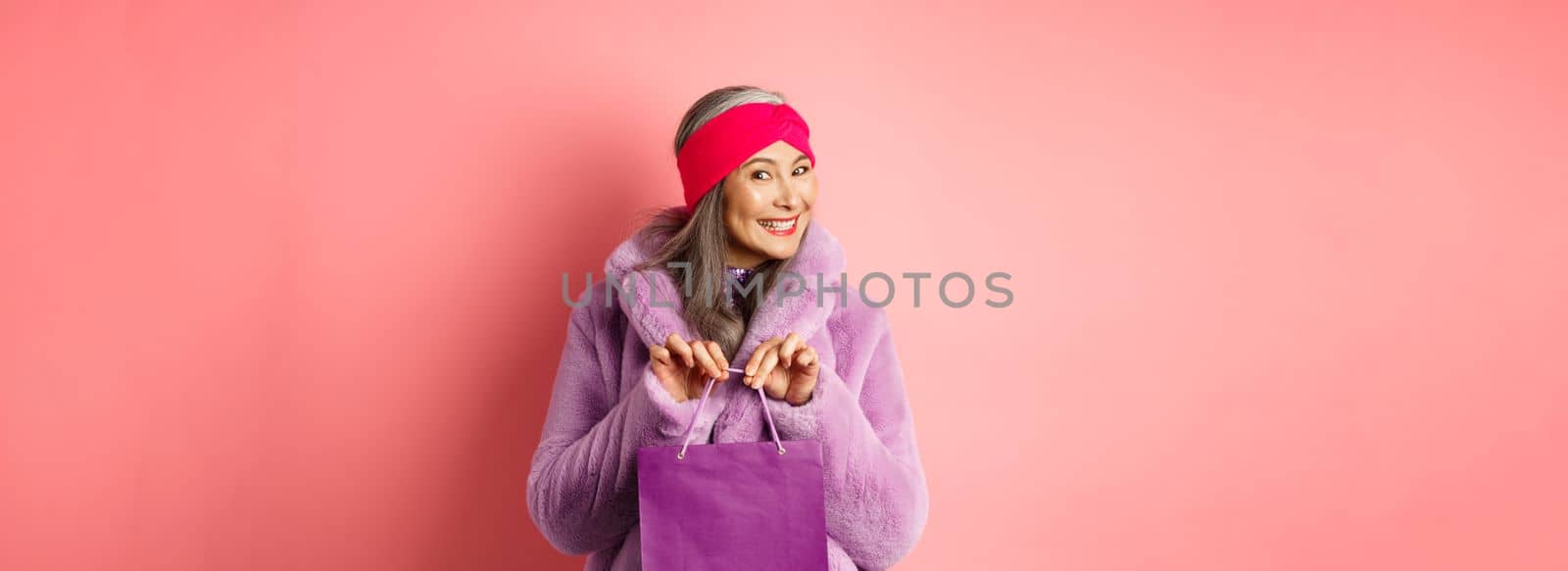 Shopping and fashion concept. Fashionable gradmother bought present and smiling, holding purple bag in hands, standing over pink background.
