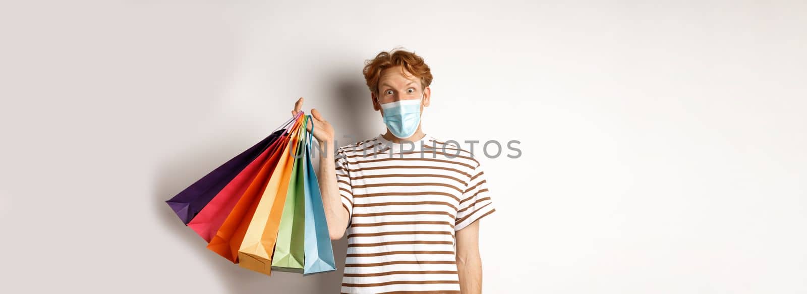 Pandemic and lifestyle concept. Handsome young man in face mask amazed with special discounts, holding shopping bags and staring excited, white background.
