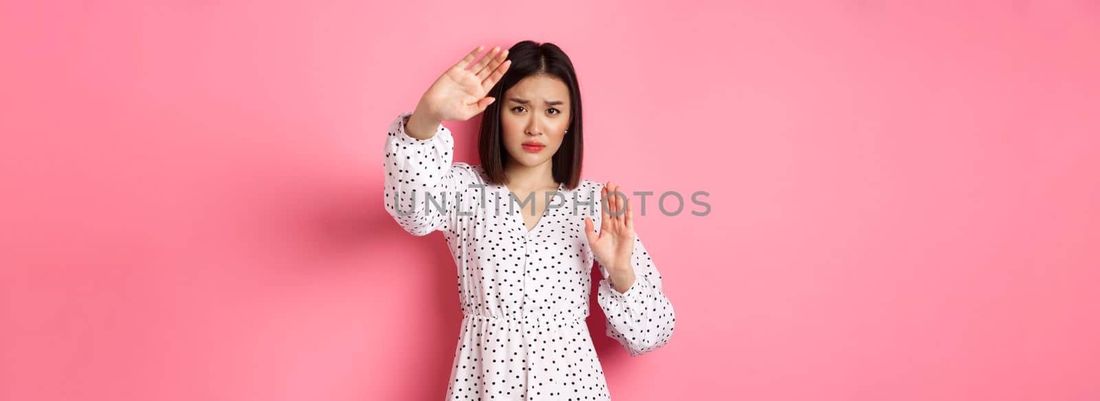 Timid and scared asian woman defending herself, raising arms in protection, victim being attacked, standing over pink background.