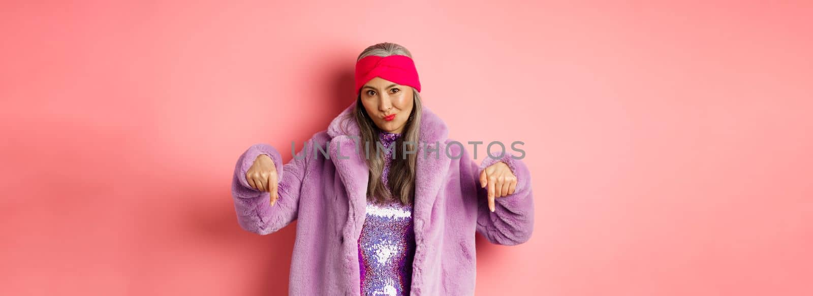 Fashion and shopping concept. Cool asian senior woman pointing fingers down, check out promo offer, looking sassy at camera, pink background.