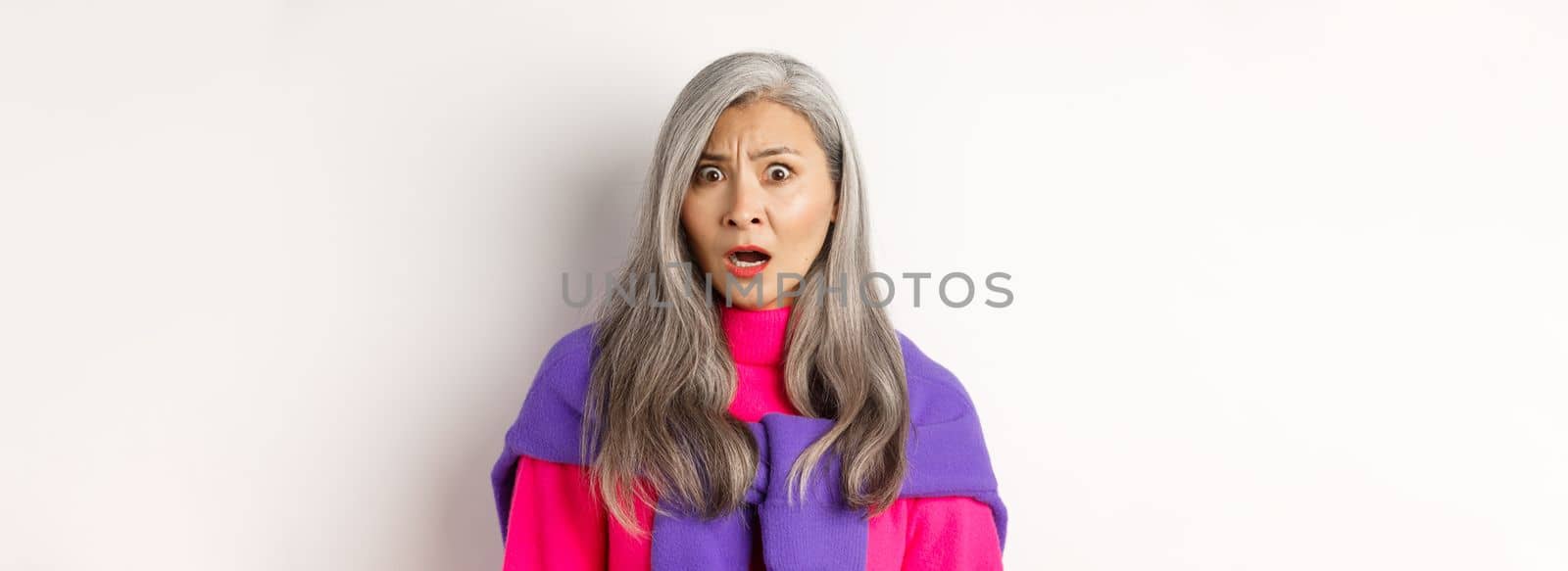 Close-up of shocked asian senior woman gasping, staring with confused face, looking with cringe, standing against white background by Benzoix